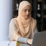 Muslim woman wearing a hijab works intently on her laptop indoors, surrounded by books.